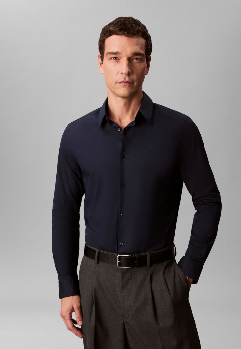 Mann mit kurzem dunklem Haar, der ein marineblaues, geknöpftes Hemd trägt, das in grau plissierte Hosen mit schwarzem Gürtel gesteckt ist, vor neutralem Hintergrund stehend.