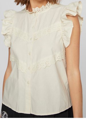 Kremowa, bezrękawnikowa bluzka z falbanami na ramionach, ozdobiona ażurową koronką na piersi i kołnierzyku, z przodu zapinana na guziki, noszona z czarnymi spodniami.