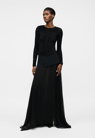 Žena v černém dlouhém topu se sklady a v černé splývavé sukni stojí s uvolněnýma rukama na světlém jednobarevném pozadí.