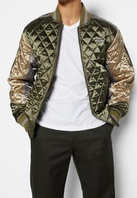 Quilted bomber jacket in olive green and beige satin, featuring ribbed cuffs and collar. Logo detail on the chest, zip-up front closure.