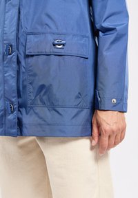 Giacca impermeabile blu con tasca frontale con chiusura a pressione e polsini con bottone, realizzata in un materiale sintetico liscio e lucido.