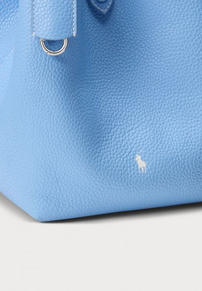 Modrá kabelka z broušené kůže s kulatým tvarem, vybavená stříbrným kováním a bílým logem poblíž spodní části. Viditelný texturovaný povrch.