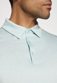 Close-up of a man wearing a light blue buttoned polo shirt with subtle geometric patterns, showing his jawline and neck.