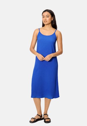 Mujer de pie con un vestido midi azul real sin mangas y sandalias planas negras frente a un fondo claro y liso.