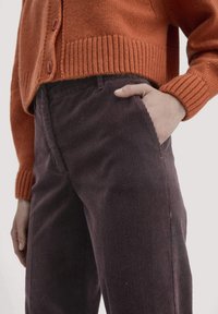 Een oranje gebreide cardigan met geribde onderrand en manchetten, gecombineerd met donkerpaarse fluwelen broek met zijzakken en verticale strepen.