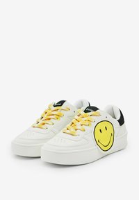 Sneakers bianchi con suola spessa, design di faccina sorridente gialla sul lato, lacci gialli con motivo e dettagli neri sul tallone.
