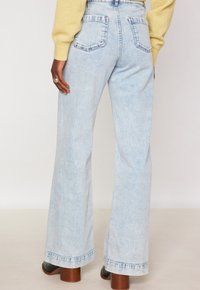 Persona che indossa jeans a vita alta larghi di un blu chiaro, maglione giallo e stivali neri con tacco, in piedi davanti a uno sfondo neutro.