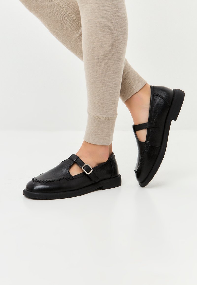 Černé kožené T-strap boty s přezkou, které mají výrazné švy a nízkou plochou podrážku. Modelka je oblečena do béžových legín.