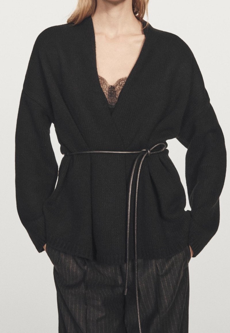 Femme portant un cardigan côtelé noir avec une fine ceinture nouée à la taille, montrant un haut à dentelle en dessous et un pantalon à fines rayures.