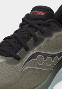 Κοντινή λήψη παπουτσιού τρεξίματος Saucony σε ελιά πράσινο και μαύρο, με κόκκινο λογότυπο στη γλώσσα και ασημένια λεπτομέρεια ρίγας στο πλάι.