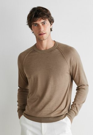 Młody mężczyzna o brązowych włosach ubrany w taupe'owy sweter z długim rękawem oraz białe spodnie, stoi na tle jednolitego jasnoszarego tła.