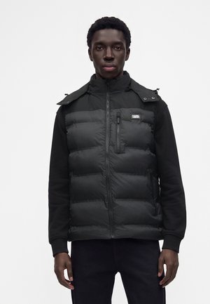 Homme portant une veste matelassée noire avec une poche poitrine zippée et un écusson logo sur une chemise noire à manches longues, debout sur un fond uni.