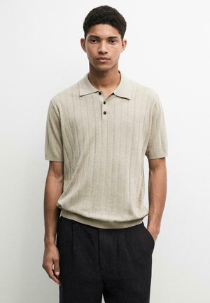 Junger Mann, der ein beiges kurzärmeliges Strickhemd mit Kragen und schwarze Hosen trägt, steht vor einem einfarbigen weißen Hintergrund.