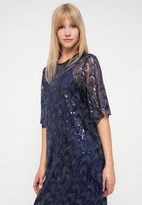 Robe bleu marine avec superposition transparente, motif floral pailleté et manches à bords festonnés. Texture lisse, design longueur genoux.