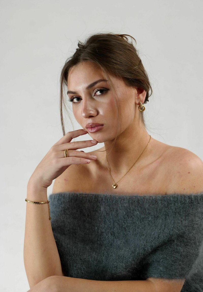 Collar de corazón de oro, pendientes de aro de oro, top de punto gris sin hombros. La textura es suave, con un brillo liso visible en la joyería.