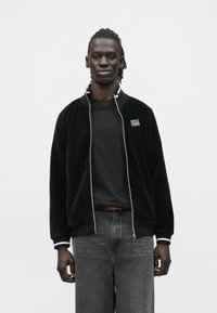 Jovem de pele escura e dreadlocks, vestindo casaco de veludo cotelê preto com acabamento branco, camiseta preta e jeans cinza, sobre fundo branco.