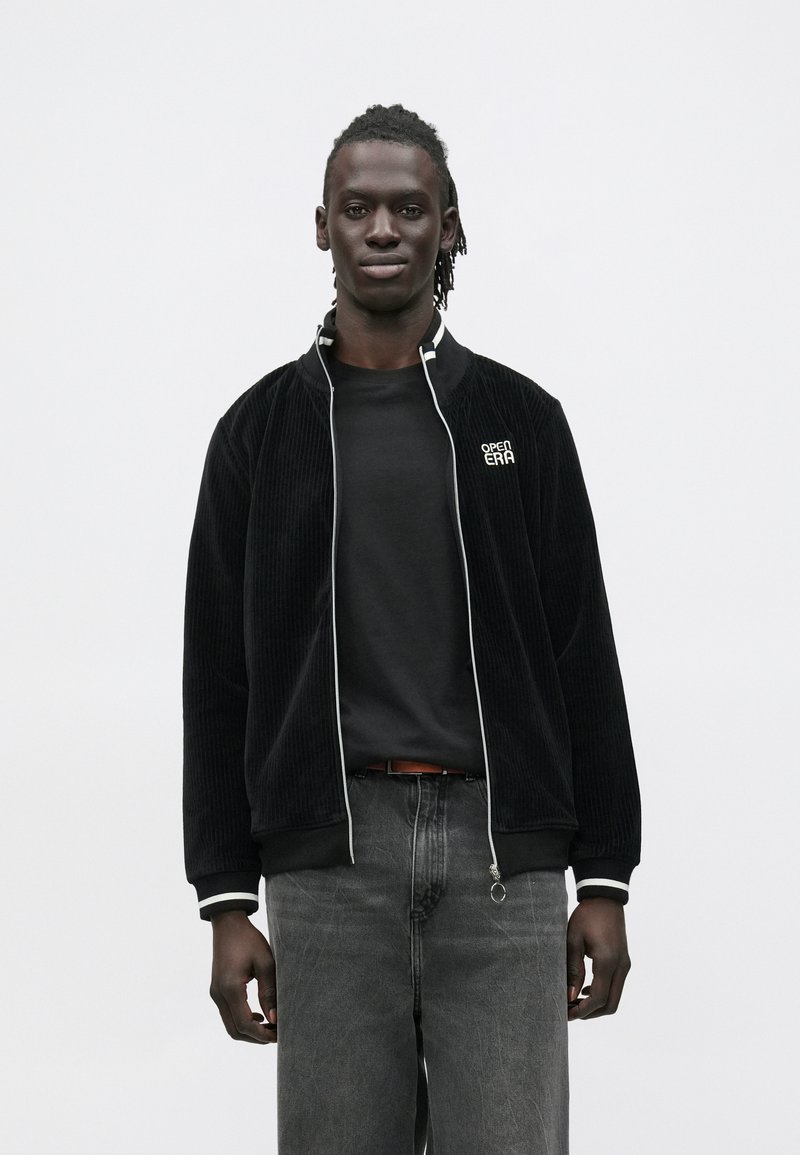 Jovem de pele escura e dreadlocks, vestindo casaco de veludo cotelê preto com acabamento branco, camiseta preta e jeans cinza, sobre fundo branco.