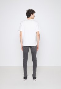 Fehér pamut póló, kerek nyakkivágással, sötétszürke, slim fit farmerrel párosítva. A modell háttal áll, fekete sportcipőt visel, fehér talppal.
