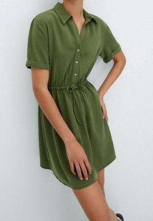 Persoon draagt een olijfgroene jurk met korte mouwen, knopen, kraag en een trekkoord in de taille, staand met één hand op de heup.