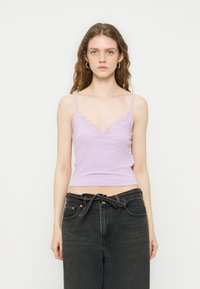 Νεαρή γυναίκα με κυματιστά μαλλιά φοράει λιλά καμιζόλα με δαντέλα και μαύρο τζιν με ψηλή μέση και δεμένη ζώνη, στέκεται μπροστά σε λευκό φόντο.