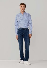 Camisa de botones en azul claro con rayas blancas, combinada con jeans oscuros y zapatillas blancas. Diseño casual y ajustado.