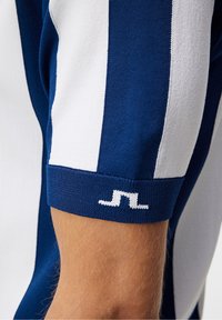 Námorně modré a bílé pruhované tričko s krátkým rukávem, s žebrovaným námořně modrým lemem a bílým logem. Tkanina se zdá být hladká a mírně texturovaná.