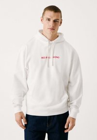 Sudadera blanca con el logo de "RED BULL RACING" en rojo, capucha con cordón, bolsillo frontal y puños de canalé. Tejido suave, corte holgado.