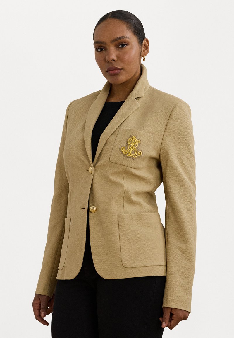 Blazer beige con un design sartorial, revers a punta, bottoni dorati, due tasche frontali e un logo ricamato sulla tasca sinistra.
