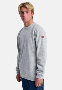 Sudadera de algodón gris con mangas largas, cuello redondo y un acabado texturizado. Presenta un pequeño parche de logo de marca marrón en el hombro izquierdo.