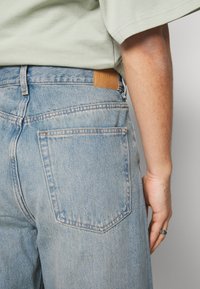 Vue de dos d'une personne portant un jean en denim bleu clair avec un patch de marque en cuir et une chemise à manches courtes vert clair, main reposant le long du corps.