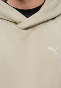 Μπεζ φούτερ με κουκούλα και λευκό λογότυπο Puma στο στήθος, φορεμένο πάνω από μια μαύρη μπλούζα, που δείχνει τον λαιμό και την περιοχή πάνω από το στήθος.