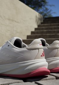 Białe sneakersy z teksturowaną siateczką na cholewce, wyściółką wokół kostki oraz różowymi akcentami na podeszwie i logo. Charakteryzują się lekką konstrukcją i zaokrąglonym kształtem.