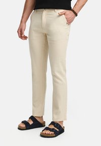 Beige chinos with a slim fit, made of lightweight fabric, featuring a waistband with belt loops and side pockets. Paired with black sandals.