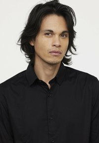 Chemise noire à boutons avec col, présentant une texture lisse et une coupe ajustée, portée par une personne ayant les cheveux foncés sur un fond neutre.