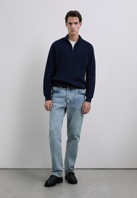 Marineblauwe geribde trui met halve ritskraag, lichtblauwe spijkerbroek en zwarte schoenen. Model staat tegen een neutrale achtergrond.