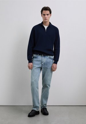 Suéter azul marino acanalado con cuello de media cremallera, jeans azul claro y zapatos negros. La modelo está de pie frente a un fondo neutro.