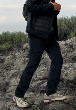 Osoba ubrana w czarną odzież outdoorową i beżowe buty trekkingowe stoi na skalistym terenie, w tle zielone krzewy.