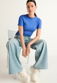 Modrá textúrovaná blúzka s krátkym rukávom skombinovaná so svetlomodrými širokými džínsami a robustnými bielymi teniskami; model sedí na priehľadnej stoličke.