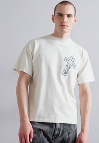 Lys beige kortermet t-skjorte laget av bomull, med et grått grafisk design foran som ligner et kors med textelementer.