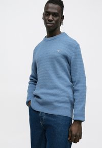 Zils faktūras džemperis ar apaļu izgriezumu, ribotu aproču un apakšmalu. Kombinēts ar tumši zilām džinsa biksēm.