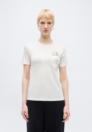 Kobieta o krótkich blond włosach, ubrana w biały t-shirt z kwiatowym wzorem na kieszeni i napisem "LOVE", w połączeniu z czarnymi spodniami.