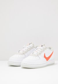 Vita sneakers med en slät läderöverdel, grå detaljer och en knallorange swoosh. Har snörning och perforerad tåsida för bättre andningsförmåga.