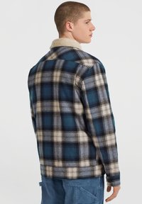 Young man facing sideways wearing a blue and beige plaid jacket with a beige sherpa collar and blue pants with a side pocket.