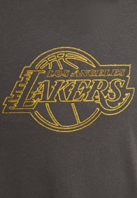 Logotipo de Los Angeles Lakers bordado en oro, que incluye el contorno del baloncesto y el texto, sobre un fondo de tela gris oscuro.