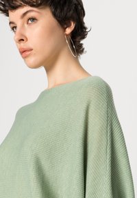 Groene ribgebreide top met een bootnek, soepel vallende mouwen en een zachte textuur, aangevuld met een grote zilveren oorbel.