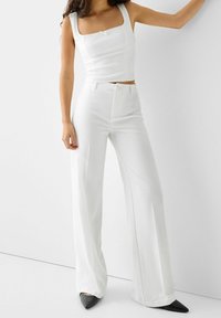 Top corto bianco aderente con scollatura quadrata e fiocco decorativo, abbinato a pantaloni bianchi a vita alta e gamba larga e scarpe con tacco a punta nere.