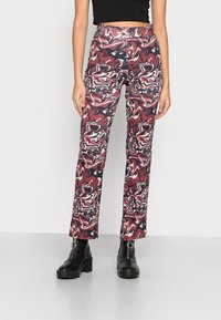 Calças ajustadas de cintura alta com um padrão abstrato em vermelho, preto e creme. Tecido suave, design de perna larga, combinadas com botas de tornozelo pretas.