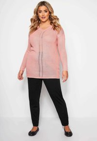 Vaaleanpunainen neulottu pitkähihaisella paidalla, jossa on V-kaula-aukko ja pystysuora raitakuosi, yhdistettynä mustiin housuihin ja matala kenkiin.