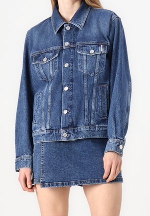 Femme portant une veste en jean bleue avec des boutons argentés, associée à une mini-jupe en jean bleue assortie sur un fond uni.