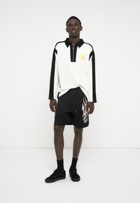 Camisa de mangas longas branca e preta com gola de fecho de meio; calções pretos com padrão tribal branco; usada com ténis pretos e meias.